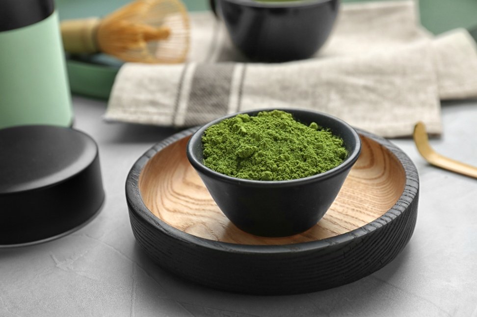 Schwarze Schale mit leuchtend grünem zeremoniellen Matcha-Pulver auf einem Holzständer, im Hintergrund sind Utensilien für die Teezeremonie zu sehen