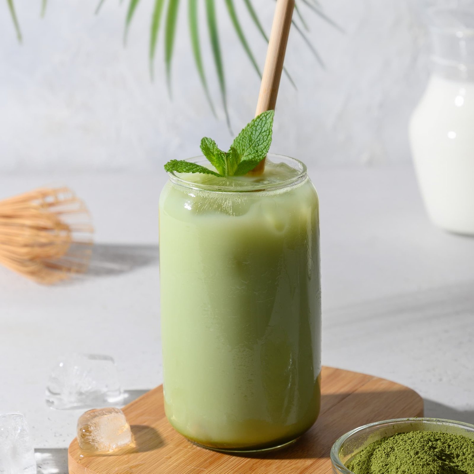 Iced matcha latte in a glass jar with a paper straw and fresh mint leaves, beside a bamboo whisk and bowl of matcha powder