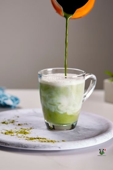 Matcha being poured from a pitcher into a glass cup with milk foam on a white saucer