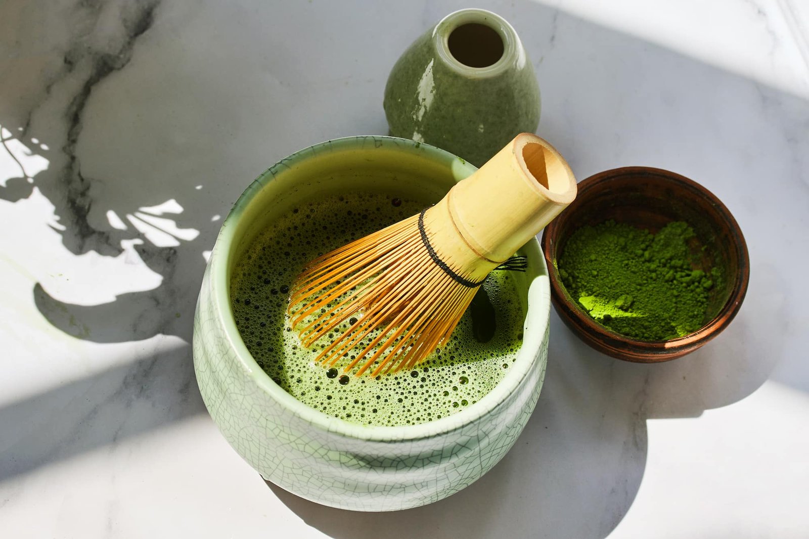 Bamboo chasen whisk in a crackled celadon ceremonial matcha bowl with foam, beside a bowl of matcha powder