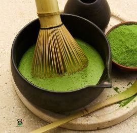 Bamboo chasen whisk in a black bowl of frothy ceremonial matcha tea with a bowl of matcha powder behind